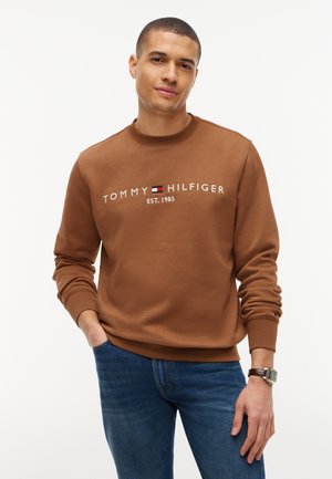 Mężczyzna ubrany w brązową bluzę Tommy Hilfiger z logo oraz ciemnoniebieskie dżinsy, stoi z jedną ręką w kieszeni na białym tle.
