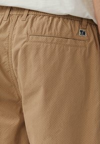 Beige gemusterte Shorts mit einem Diamantdesign, elastischem Bund und einer einzelnen Gesäßtasche. Die Textur wirkt glatt und leicht.