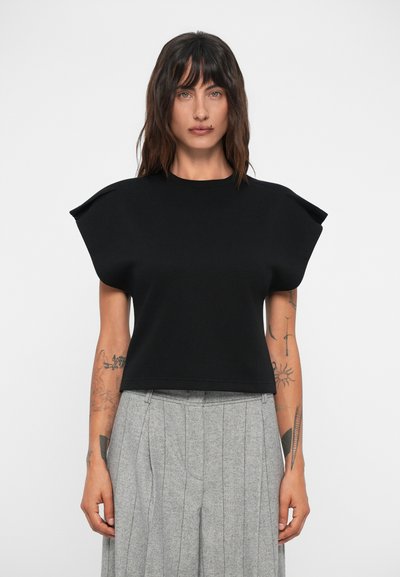 Top corto nero con design strutturato sulle spalle, maniche corte e una texture morbida. Abbinato a pantaloni plissettati grigio chiaro.