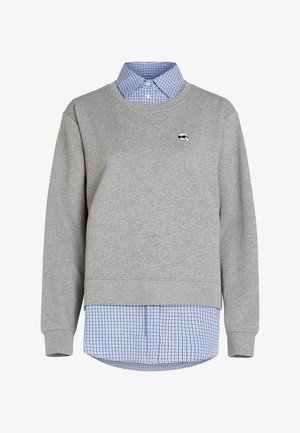 Szara bluza z okrągłym dekoltem założona na niebieską i białą koszulę w kratę z guzikami, z małym haftowanym logo na lewej piersi.