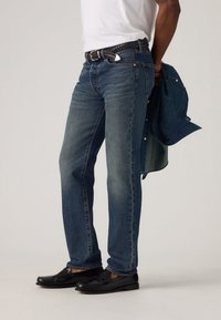 Σκούρο μπλε τζιν με κλασική εφαρμογή, συνδυασμένο με λευκό t-shirt και μαύρα μοκασίνια. Ένα τζιν μπουφάν είναι περασμένο στο χέρι.