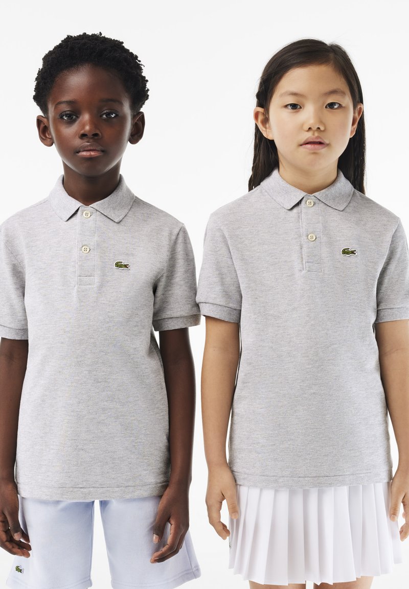 Due bambini indossano polo grigie a maniche corte con bottoni. Ogni maglietta presenta il logo Lacoste. Un bambino indossa un pantalone corto bianco, l'altro una gonna bianca plissettata.