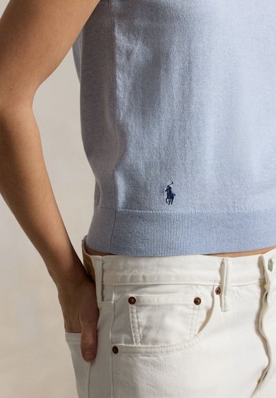 Άτομο που φοράει ανοιχτό μπλε αμάνικο μπλουζάκι με μικρό ναυτικό λογότυπο παίκτη πόλο, ελαφρώς μπουκαδωμένο σε ψηλόμεσο λευκό τζιν με το χέρι στην τσέπη.