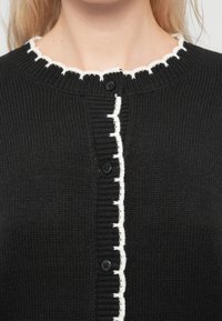 Μαύρη πλεκτή ζακέτα με υφή, που διαθέτει αντίθετη λευκή διακόσμηση κατά μήκος του στρογγυλού γιακά και της λωρίδας με κουμπιά.