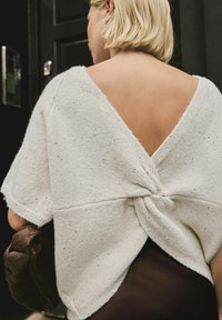 Woman with short blonde hair wearing white textured top with open back twist detail, holding a brown woven handbag against dark background.