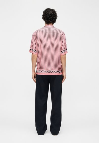 Άνδρας με σγουρά μαλλιά, που φοράει ροζ πουκάμισο με κοντά μανίκια και μαύρα σχέδια καρδιών, καθώς και μαύρο παντελόνι, στέκεται με την πλάτη σε απλό φόντο.