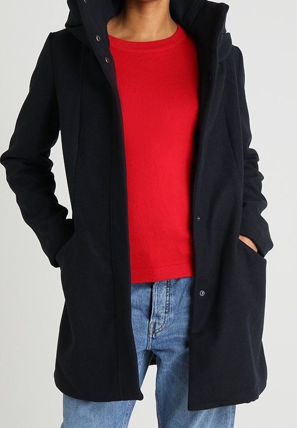 Persona che indossa un cappotto nero con cappuccio sopra un maglione rosso e jeans blu, con le mani nelle tasche del cappotto.