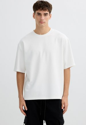 Joven con cabello corto y rizado, vistiendo una camiseta blanca grande y pantalones cargo negros, de pie frente a un fondo blanco.