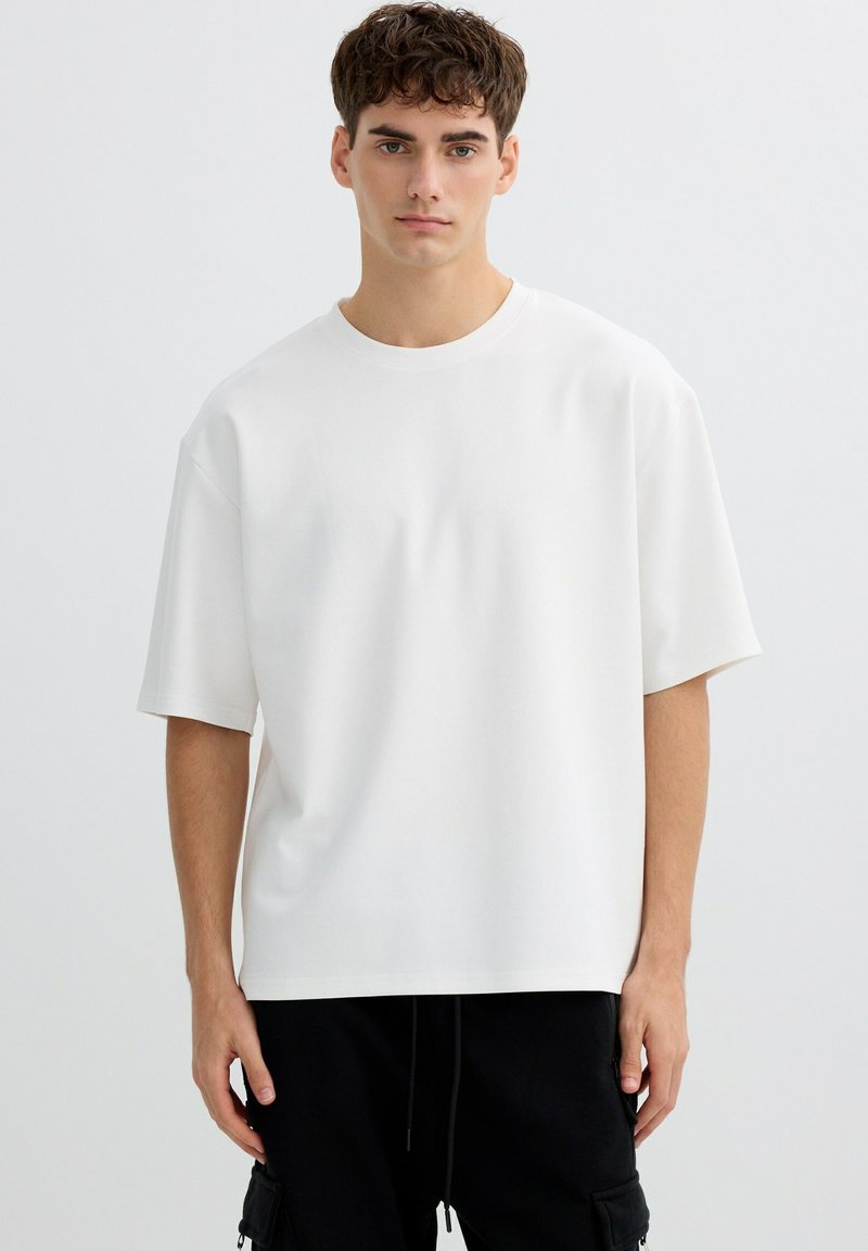 Joven con cabello corto y rizado, vistiendo una camiseta blanca grande y pantalones cargo negros, de pie frente a un fondo blanco.