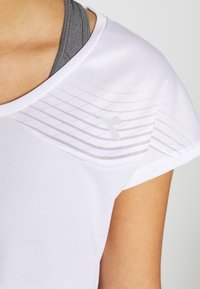 Bílý sportovní tričko s šedým geometrickým vzorem na rameni, nošené přes šedou sportovní podprsenku, viditelné na horní části trupu osoby.