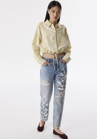Geel linnen cropped shirt met trekkoord in de taille, gecombineerd met verstoorde lichtblauwe jeans voorzien van handgeschreven tekst en scheuren. Bruine ballerina's gedragen.