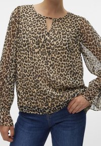 Blusa con stampa leopardata in tessuto trasparente, caratterizzata da un collo a chiave e maniche lunghe con volant. Indossata con jeans blu.