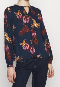 Blusa blu navy con motivi floreali e uccelli in rosso, rosa e arancione; presenta un colletto a occhiello e maniche lunghe con polsini elasticizzati.