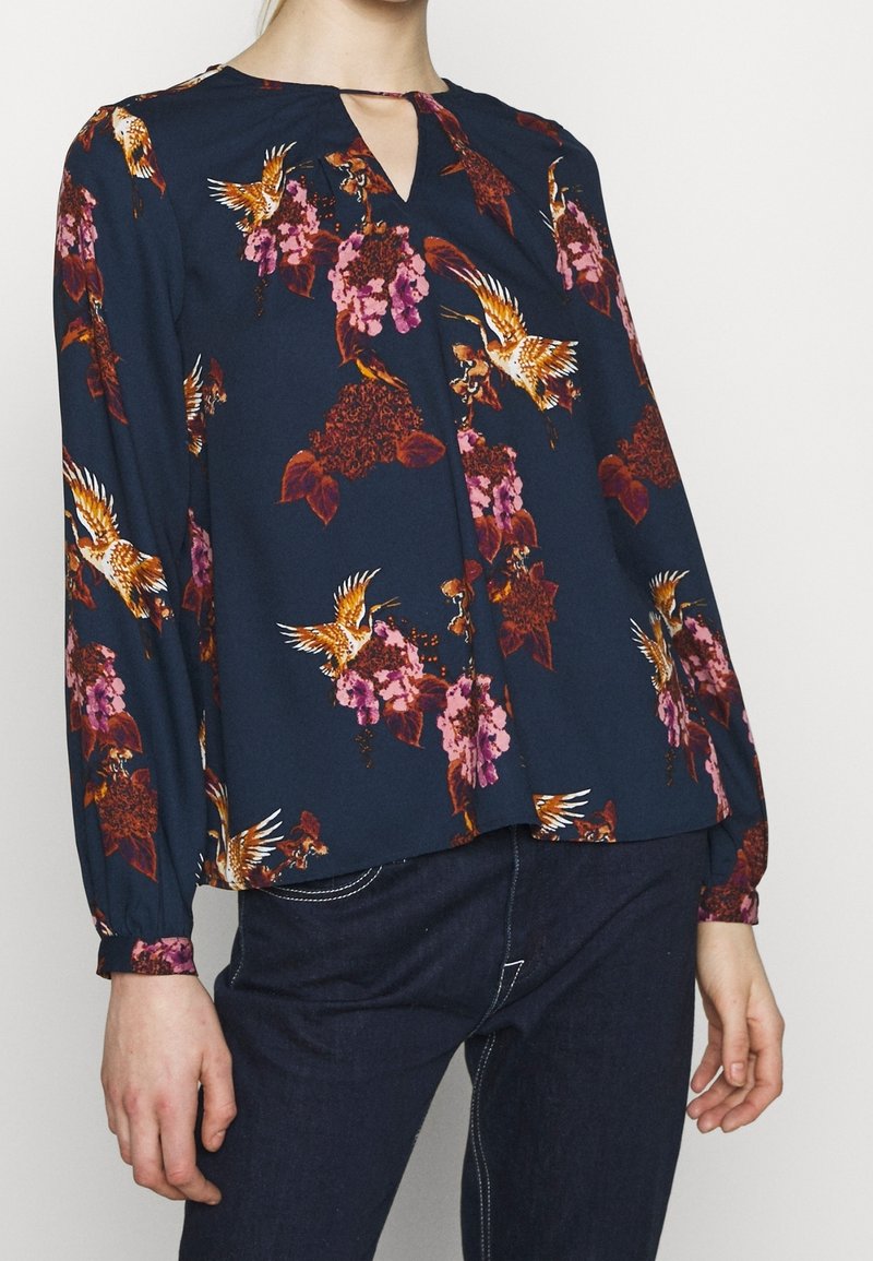 Blusa blu navy con motivi floreali e uccelli in rosso, rosa e arancione; presenta un colletto a occhiello e maniche lunghe con polsini elasticizzati.