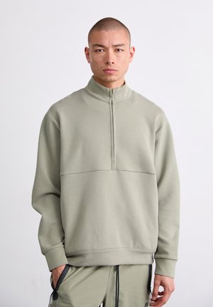 Zipped pullover in soft, light green fabric. Features a high collar and a seam across the chest. Model wears matching pants with pockets.