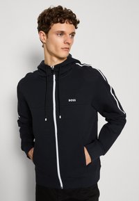 Svart zip-up hoodie med vita accentlinjer på ärmarna. Har justerbara snoddar, en zip-försklassning fram och BOSS-logotyp på bröstet.