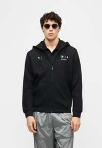 Νεαρός άνδρας που φοράει μαύρο φούτερ με φερμουάρ BMW Motorsport, μαύρα γυαλιά ηλίου και λαμπερό ασημί-γκρι παντελόνι, στέκεται μπροστά σε απλό φόντο.