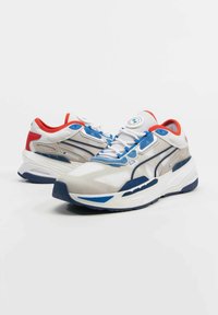 Hvite og blå sporty sneakers med teksturert mesh-overdel, røde detaljer, og en kraftig såle med blå dekorasjoner og en logo på tungen.