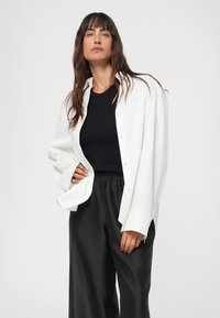 White oversized button-up shirt made of smooth fabric, paired with a black fitted top and black wide-leg trousers, showcasing a relaxed fit.