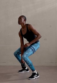 Čierny top bez rukávov, modré legíny s hadím vzorom a horizontálnymi pruhmi, drží kettlebell, čierne športové topánky, v pozícii drepu.