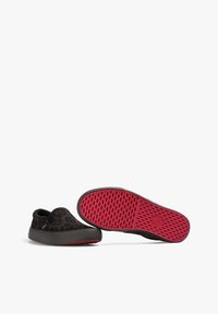 Par de zapatos negros tipo slip-on con un sutil estampado de leopardo y suelas de goma texturizada en rojo brillante, un zapato en posición vertical y el otro mostrando la suela.
