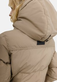 Beige dunjakke med stor, isolert hette. Har detaljer med søm og en svart logotagg bak, laget av glatt stoff.