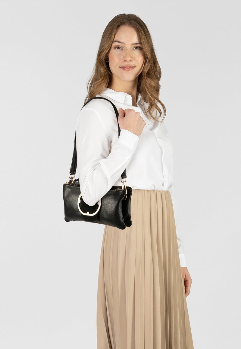 Bolso de mano de cuero negro con herrajes circulares plateados, cierre de cremallera y asas para el hombro, sostenido por una persona que lleva una camisa blanca y una falda beige.