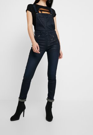 Denim overalls in dark blue with adjustable straps, a zip front, and fitted legs, paired with a black t-shirt featuring a metallic graphic design.