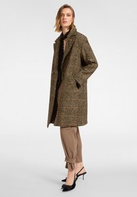 Brązowy i żółty płaszcz w kratę z wyłogami i dwoma bocznymi kieszeniami, zestawiony z beżowymi spodniami i czarnymi butami na obcasie typu slingback.