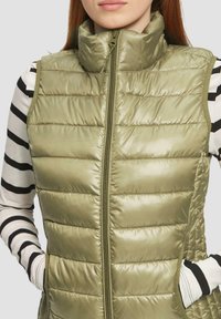 Olivgrüner Puffer-Weste mit hohem Kragen, Reißverschluss vorne und Seitentaschen. Die Nähte bilden ein gestepptes Muster, hergestellt aus glänzendem Material.