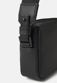 Sac à épaule noir texturé avec un connecteur de sangle robuste et des accents en métal ; présente une surface lisse et des bords nets.