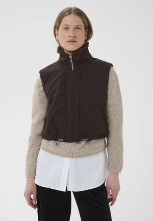 Jeune femme portant un gilet noir sans manches à fermeture éclair par-dessus un pull beige et une chemise blanche avec un pantalon noir, debout devant un arrière-plan uni.