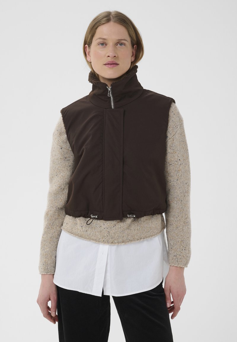 Jeune femme portant un gilet noir sans manches à fermeture éclair par-dessus un pull beige et une chemise blanche avec un pantalon noir, debout devant un arrière-plan uni.