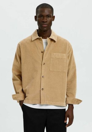 Homme portant une chemise boutonnée en velours côtelé beige par-dessus un t-shirt blanc et un pantalon noir, debout devant un fond clair uni.