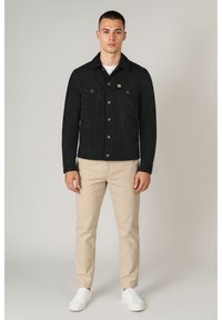 Schwarze Wildlederjacke mit zwei Brusttaschen, Knöpfen vorne und Hemdkragen. Kombiniert mit hellbeigen Hosen und weißen Turnschuhen.