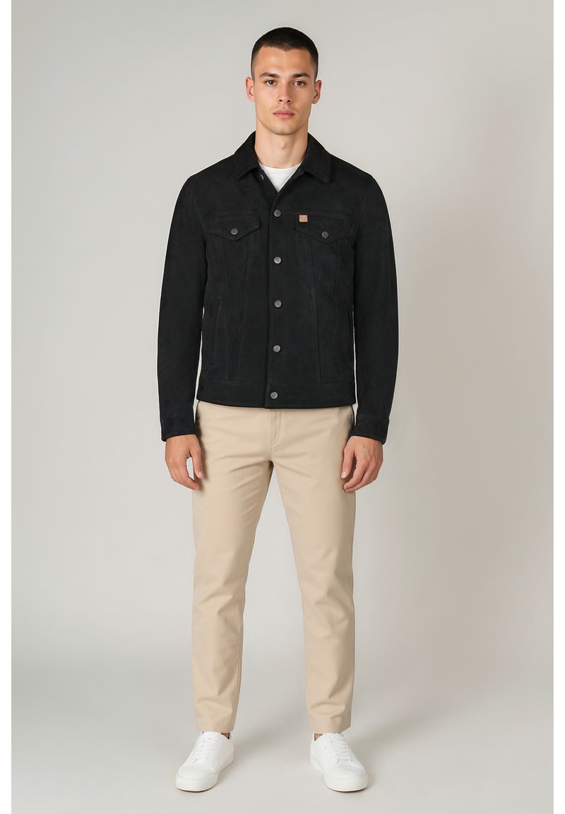 Schwarze Wildlederjacke mit zwei Brusttaschen, Knöpfen vorne und Hemdkragen. Kombiniert mit hellbeigen Hosen und weißen Turnschuhen.