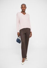 Pulover roz deschis cu decolteu în V, pantaloni maro cu model, tocuri albe și o geantă mică albastră cu un design, purtate de o persoană în picioare.