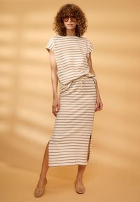 Femme aux cheveux bouclés portant de grandes lunettes de soleil et un ensemble haut et jupe beige à rayures, debout devant un fond beige.