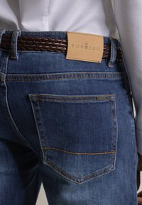 Pantalones vaqueros de mezclilla en azul oscuro con un desvanecimiento sutil, costuras detalladas y una etiqueta de cuero color canela con la inscripción "PUROEGO" visible en la cintura.