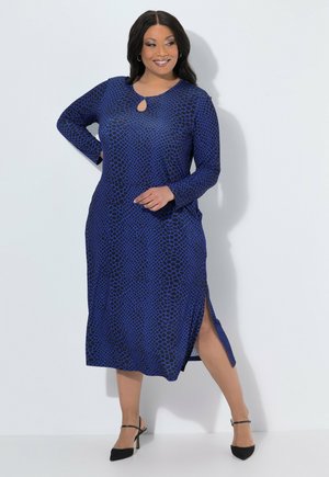Donna che indossa un vestito blu a maniche lunghe con macchie astratte nere, spacco laterale e tacchi neri con cinturino alla caviglia, posando su sfondo bianco.