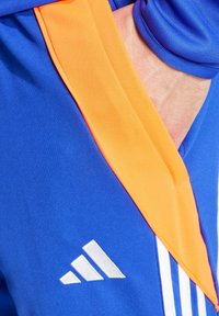 Plave sportske šortse s narančastim bočnim trakama, izrađene od teksturirane tkanine, s bijelim logotipom na rubu. Uključuju detalj bočne džepa.