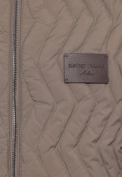 Quiltota brūna jaka ar rāvējslēdzēju, ar ševronu rakstiem un teksturētu ādas birku, uz kuras ir rakstīts "EMPORIO ARMANI."