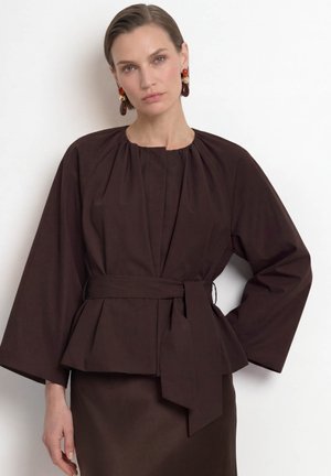 Woman wearing a dark brown belted top with wide sleeves and matching skirt, standing against a plain white background.