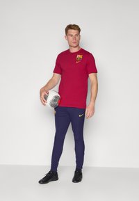 Rød kortærmet T-shirt med Barcelona-logo og gul Nike-swoosh, parret med mørkeblå ribbede bukser og sorte fodboldsko, mens han holder en bold.