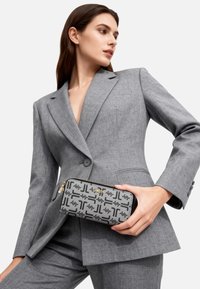 Clutch grigio con motivo triangolare e zip nera, con logo dorato. Il modello indossa un completo grigio abbinato dalla silhouette aderente.