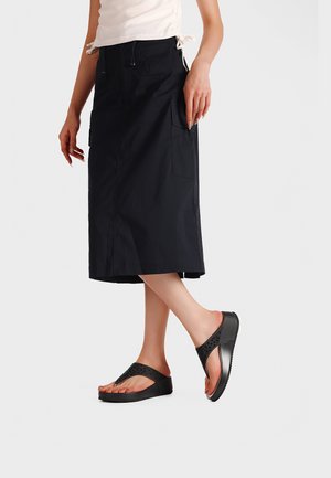 Donna che indossa una gonna midi nera con tasche laterali e sandali neri con plateau e cinturini testurizzati, in piedi con un piede leggermente sollevato.