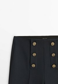 Mustat räätälöidyt housut korkealla vyötäröllä, joissa on kuusi kultaa koristepainiketta edessä ja sileä, teksturoitu kangas.