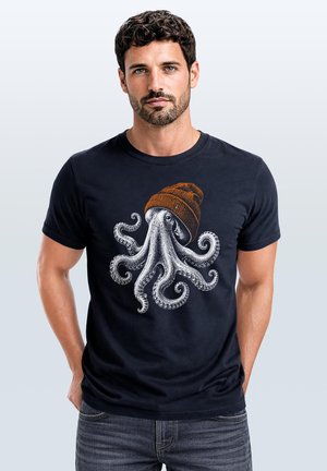 Mann mit kurzen dunklen Haaren und Bart, der ein schwarzes T-Shirt mit einem Oktopus trägt, der eine braune Strickmütze trägt.