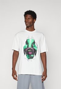Hvid bomulds-T-shirt med et farverigt grafisk design i sort og grønne nuancer, fremhævet af et Nike-logo. Korte ærmer, afslappet pasform.