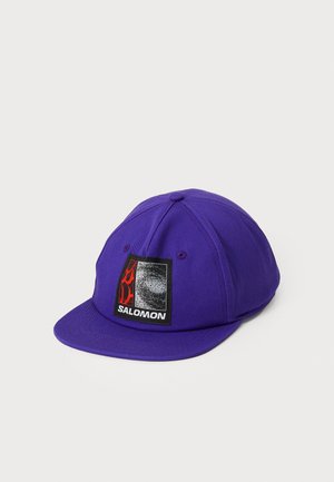 Chapéu de tecido roxo com uma aba plana, apresentando um brilho retangular preto e vermelho na frente com a inscrição "SALOMON".